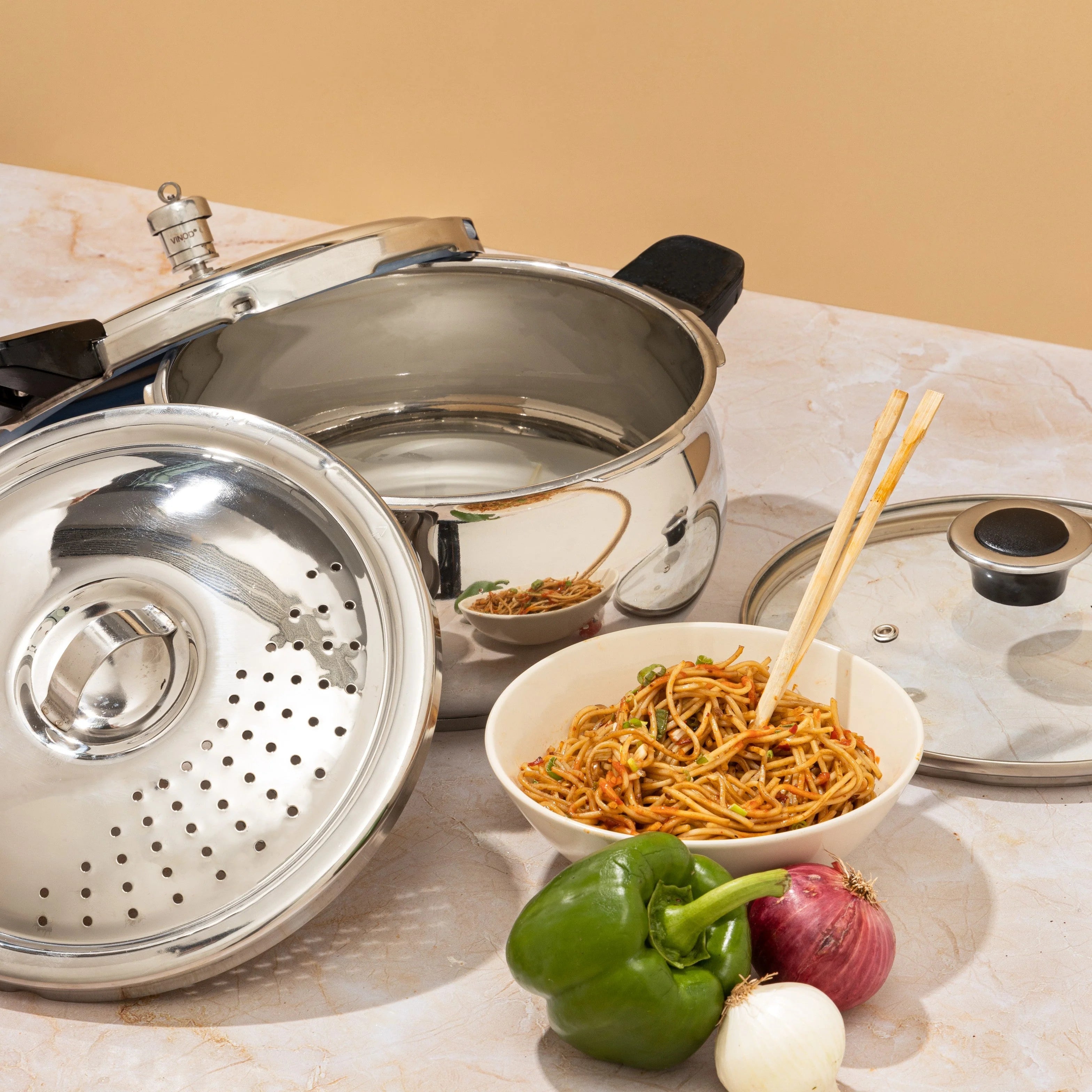5.5 liter Stainless steel Pressure Cooker set with strainer and Lid along with a bowl of noodles and vegetables on a marble surface.