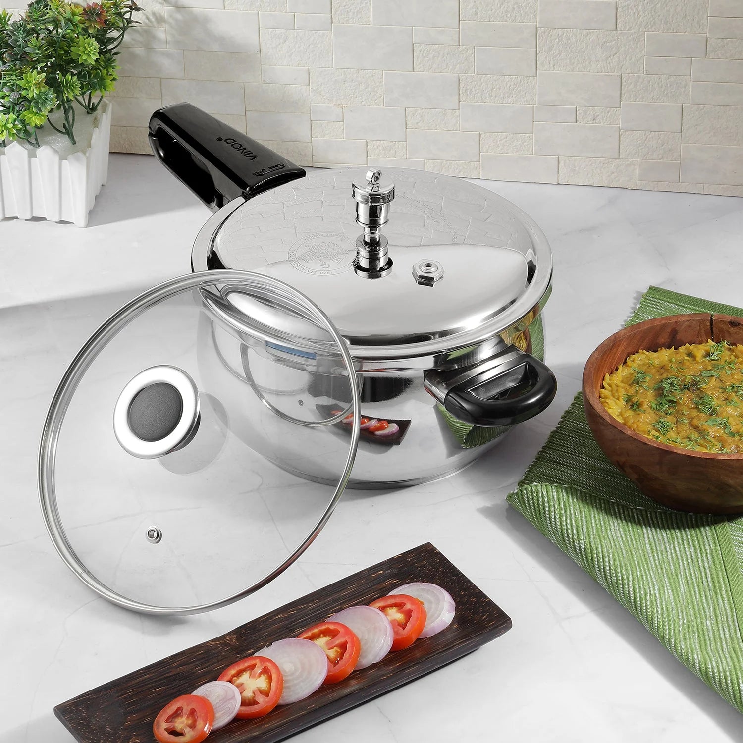 6.5 liters Stainless steel pressure cooker with glass lid on a kitchen counter, surrounded by vegetables and a bowl of food.