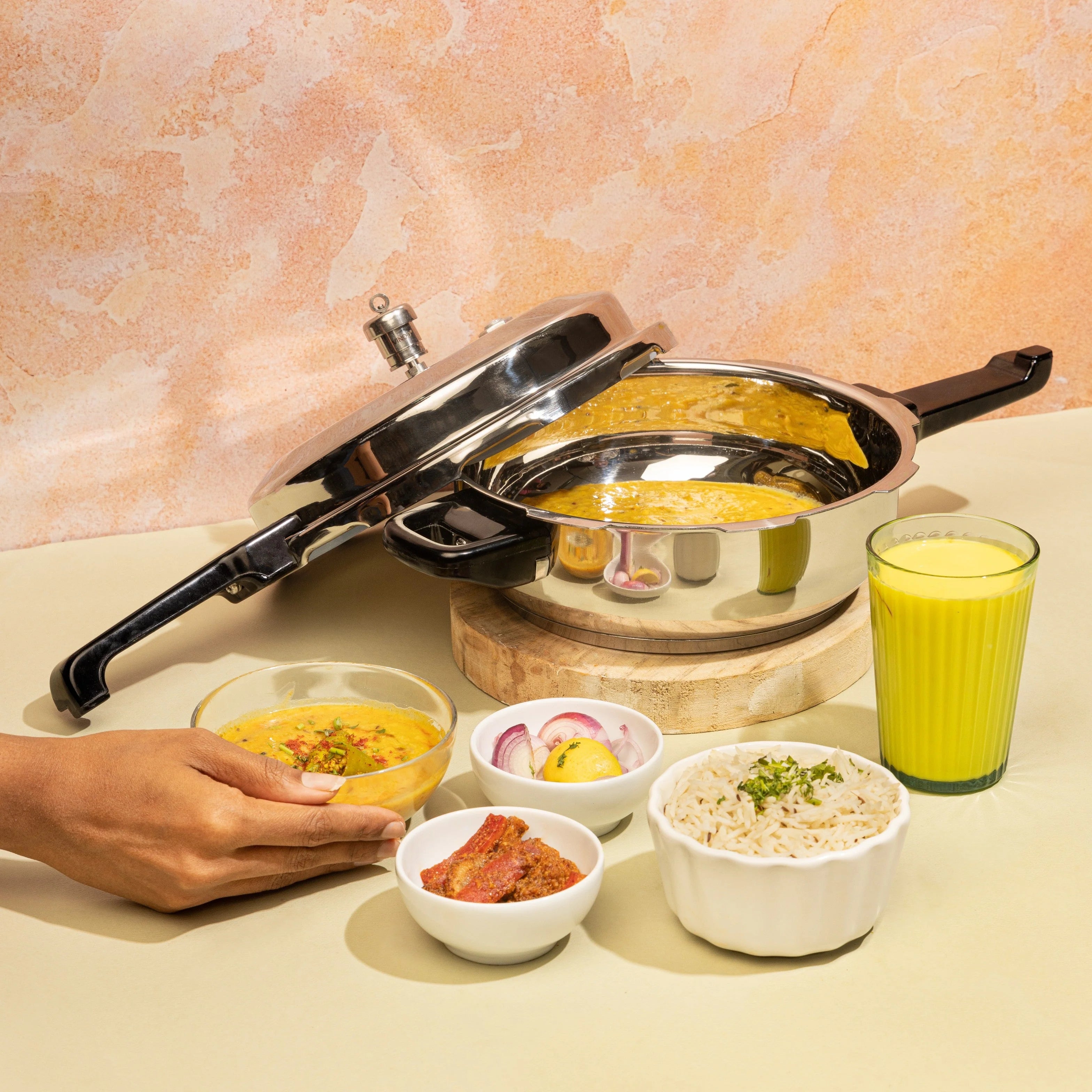 Pressure cooker with a lid open, showing food inside, on a kitchen counter with various dishes and a glass of juice.