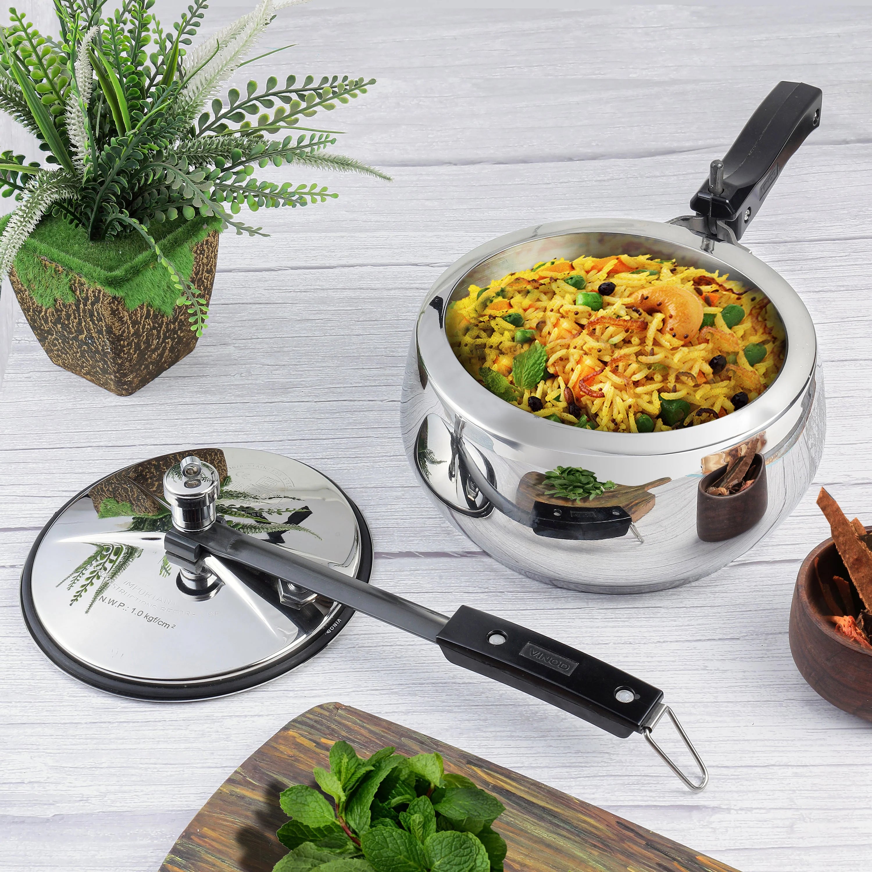 3.5 liters Stainless steel pressure cooker with lid on a light wooden surface, surrounded by greenery.