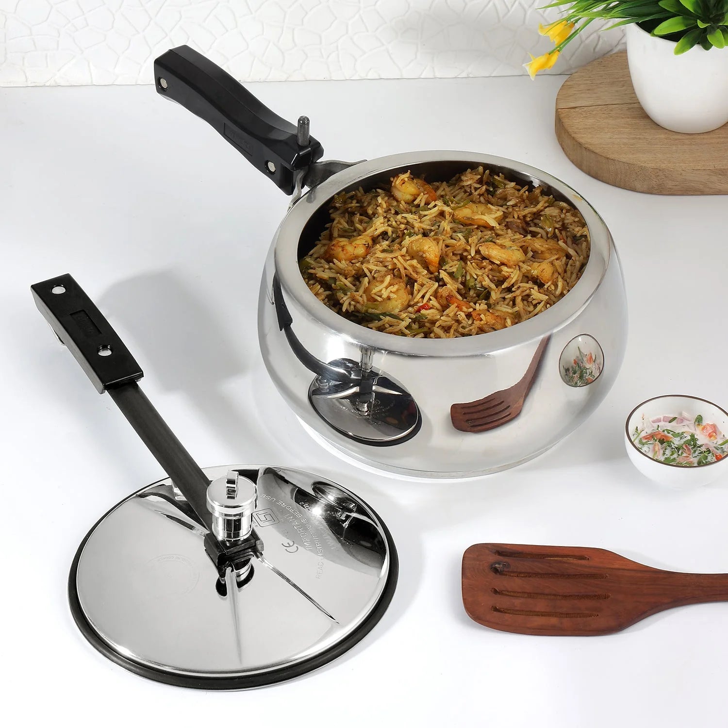 5 Liters Stainless steel pressure cooker with lid on a white surface, surrounded by a wooden spatula and small dish.