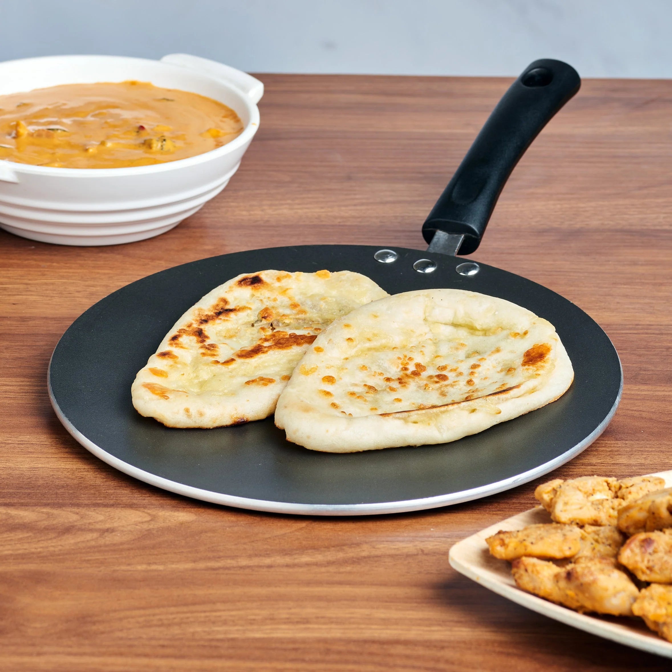 Two pieces of naan bread on a black Tawa with a bowl of curry and a plate of fried chicken on a wooden table.
